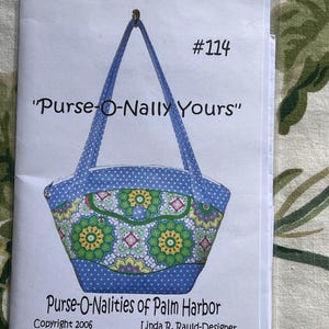 May include: A blue and white polka dot purse with a green and yellow floral pattern. The purse has a flap with a pocket and a blue button. The text on the image reads "#114 "Purse-O-Nally Yours" Purse-O-Nalities of Palm Harbor Copyright 2006 Linda R. Rauld-Designer".