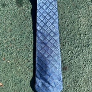 May include: A blue necktie with a repeating diamond pattern. The tie is a medium shade of blue and has a subtle sheen. The diamond pattern is slightly darker than the base color, creating a textured effect. The tie is lying flat on a green surface.