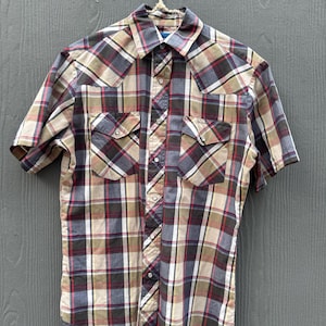 May include: A short-sleeved plaid shirt in shades of brown, red, blue, and beige hangs from a hanger. The shirt features two chest pockets and a button-down front. The plaid pattern is a classic design.