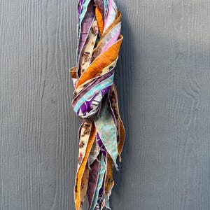 May include: A colorful scarf made of various patterned fabric strips. The scarf features a mix of orange, purple, blue, and cream colors. The scarf is knotted at the top and hangs vertically, showcasing the different patterns and textures.