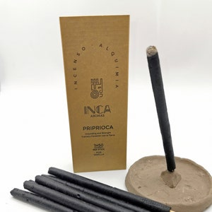 May include: Image of Inca Aromas incense sticks. Several black incense sticks are visible, along with a lit stick in a clay holder. A cardboard box displays the brand name and product details, including the text "PRIPRIOCA".