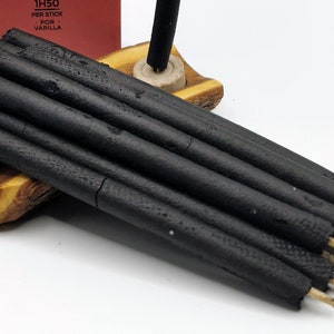 Puede incluir: Un paquete de varillas de incienso negras con puntas de madera, descansando sobre un soporte de madera. Las varillas est&aacute;n dispuestas en forma de abanico. El embalaje del incienso es visible en el fondo, con el texto "1H50 PER STICK POR VANILLA".