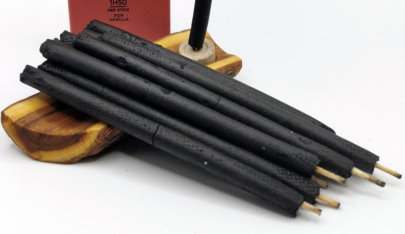 Puede incluir: Un paquete de varillas de incienso negras con puntas de madera, descansando sobre un soporte de madera. Las varillas est&aacute;n dispuestas en forma de abanico. El embalaje del incienso es visible en el fondo, con el texto "1H50 PER STICK POR VANILLA".