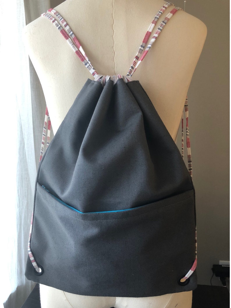 May include: A gray drawstring backpack with a zippered pocket and red and white plaid drawstrings.