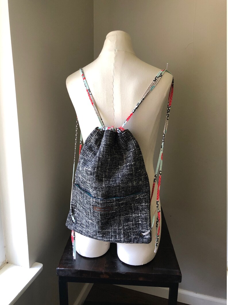 May include: Black and white drawstring backpack with a small pocket on the front. The straps are made of a colorful fabric with red, white, and blue accents.