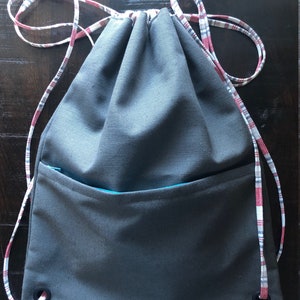 May include: A gray drawstring bag with a red and white plaid drawstring and a teal zippered pocket. The bag has two grommets on the bottom.