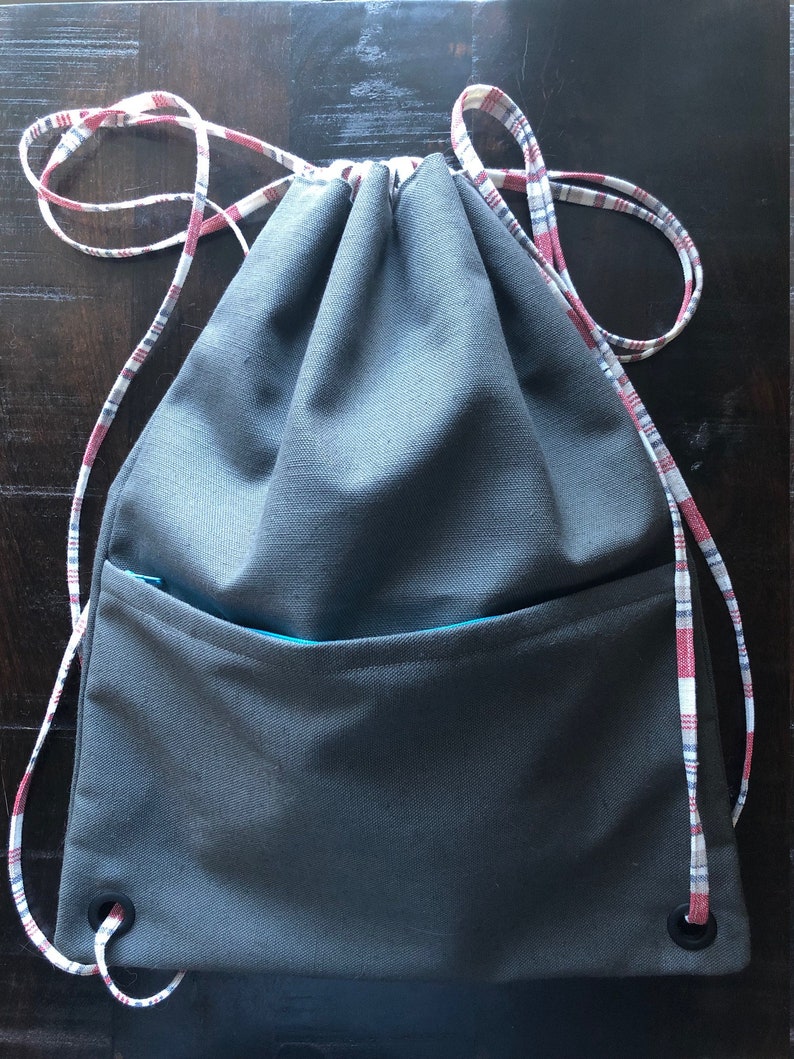 May include: A gray drawstring bag with a red and white plaid drawstring and a teal zippered pocket. The bag has two grommets on the bottom.