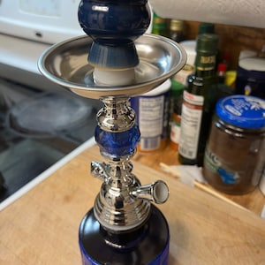 May include: A silver and blue hookah with a blue glass base and a silver bowl. The hookah is on a wooden surface.