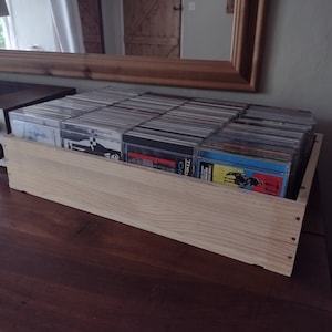 Puede incluir: Una caja de madera llena de CD, colocada sobre una superficie de madera oscura. La caja es de madera clara y está llena de varias carcasas de CD. Un receptor estéreo es visible a la izquierda, y un espejo cuelga en el fondo.
