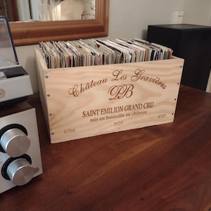 Puede incluir: Una caja de madera con la inscripción "Château Les Gravières" llena de discos de vinilo. La caja tiene texto grabado y está sobre una superficie de madera. En el fondo se ven un equipo de música y un tocadiscos vintage.