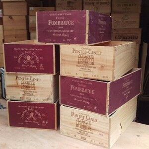 Pu&ograve; includere: Una pila di sei casse di legno per vino con etichette bordeaux. Le etichette mostrano i nomi di diversi vini, tra cui Ch&acirc;teau Pape Cl&eacute;ment, Pontet-Canet e Fombrauge.
