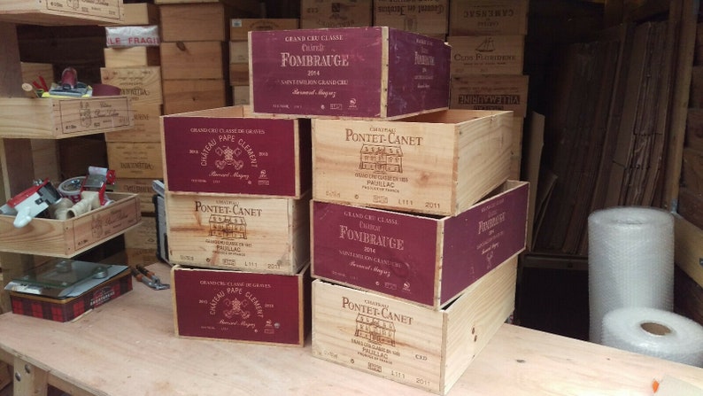 Pu&ograve; includere: Una pila di sei casse di legno per vino con etichette bordeaux. Le etichette mostrano i nomi di diversi vini, tra cui Ch&acirc;teau Pape Cl&eacute;ment, Pontet-Canet e Fombrauge.