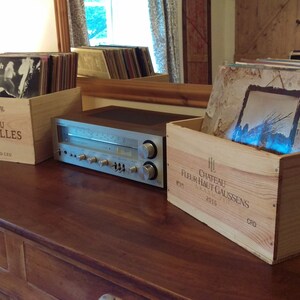 Pu&ograve; includere: Due casse di legno per vino, una etichettata "Chateau Grenouilles" e l'altra "Chateau Haut Caussens", sono utilizzate per riporre dischi in vinile. Un ricevitore stereo argentato si trova su un tavolo di legno tra le casse.