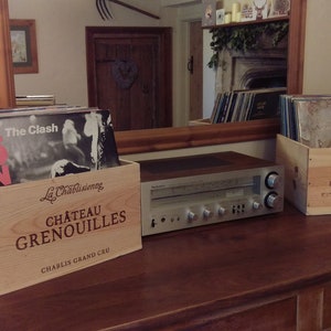 Pu&ograve; includere: Due casse di legno per vino, una etichettata "Chateau Grenouilles Chablis Grand Cru" e l'altra "Chateau Fleur Haut Gaussens 2018", sono impilate con dischi in vinile. Un ricevitore stereo vintage si trova su un tavolo di legno.