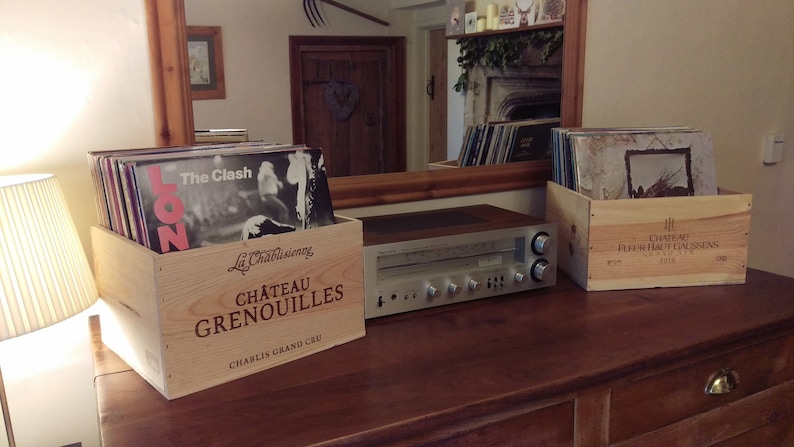 Pu&ograve; includere: Due casse di legno per vino, una etichettata "Chateau Grenouilles Chablis Grand Cru" e l'altra "Chateau Fleur Haut Gaussens 2018", sono impilate con dischi in vinile. Un ricevitore stereo vintage si trova su un tavolo di legno.