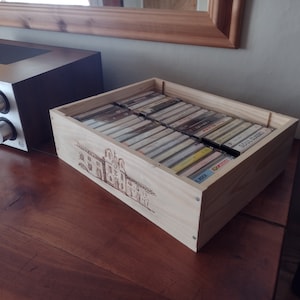 May include: A wooden box with a burnt-in design of a building, filled with cassette tapes. The box is sitting on a wooden surface.