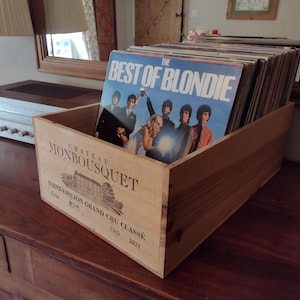 May include: A rectangular wooden crate featuring the printed text Chateau Monbousquet Saint-Emilion Grand Cru Classe 2021, filled with a collection of vinyl records. The front-most record sleeve displays the title The Best of Blondie.