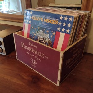 Pu&ograve; includere: Una scatola di legno con un'etichetta rossa e bianca che dice "Chateau Fombrauge 2013 Bernard Magrez". La scatola &egrave; piena di dischi in vinile, tra cui uno con un design di bandiera americana rosso, bianco e blu e il testo "Kelly's Heroes".