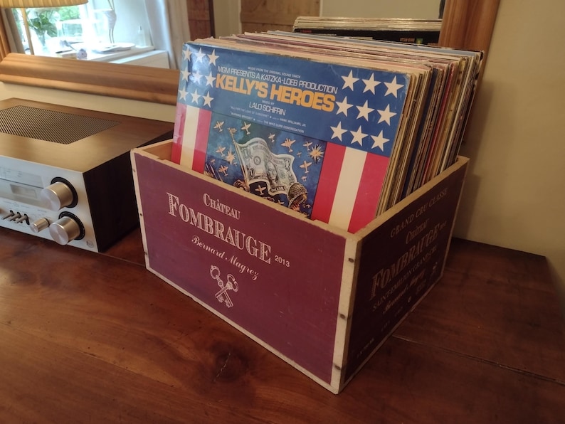 Pu&ograve; includere: Una scatola di legno con un'etichetta rossa e bianca che dice "Chateau Fombrauge 2013 Bernard Magrez". La scatola &egrave; piena di dischi in vinile, tra cui uno con un design di bandiera americana rosso, bianco e blu e il testo "Kelly's Heroes".