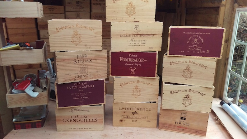 Pu&ograve; includere: Una pila di casse di vino in legno con diverse etichette. Le casse sono impilate a forma di piramide e sono in legno marrone chiaro. Le etichette sono stampate in bianco e nero e presentano i nomi di diversi vini. Alcune etichette sono in francese, mentre altre sono in inglese.