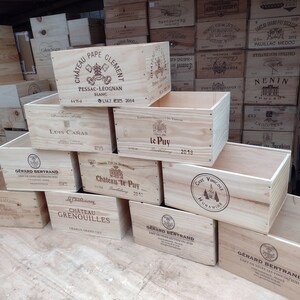 Pu&ograve; includere: Una pila di casse di legno per vino con varie etichette, tra cui Chateau Pape Clement, Luis Ca&ntilde;as, Chateau te Puy e Gerard Bertrand. Le casse sono in legno naturale e hanno un aspetto rustico.