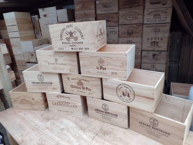 Pu&ograve; includere: Una pila di casse di legno per vino con varie etichette, tra cui Chateau Pape Clement, Luis Ca&ntilde;as, Chateau te Puy e Gerard Bertrand. Le casse sono in legno naturale e hanno un aspetto rustico.