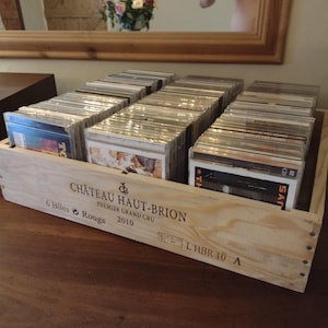 Könnte beinhalten: Eine Holzkiste mit dem Etikett "Château Haut-Brion Premier Grand Cru 2010" und dem Text "6 Biles Rouge" und "LHBR 10 A". Die Kiste ist mit einer Sammlung von CDs gefüllt.