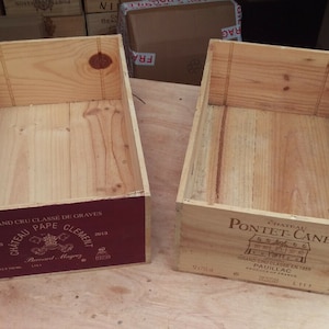 Pu&ograve; includere: Due cassette di legno per vino vuote. La cassetta a sinistra ha un'etichetta rossa con il testo "Grand Cru Classe de Graves Chateau Pape Clement 2012." La cassetta a destra ha un'etichetta bianca con il testo "Chateau Pontet-Canet Grand Cru Classe 1855 Pauillac 2011."