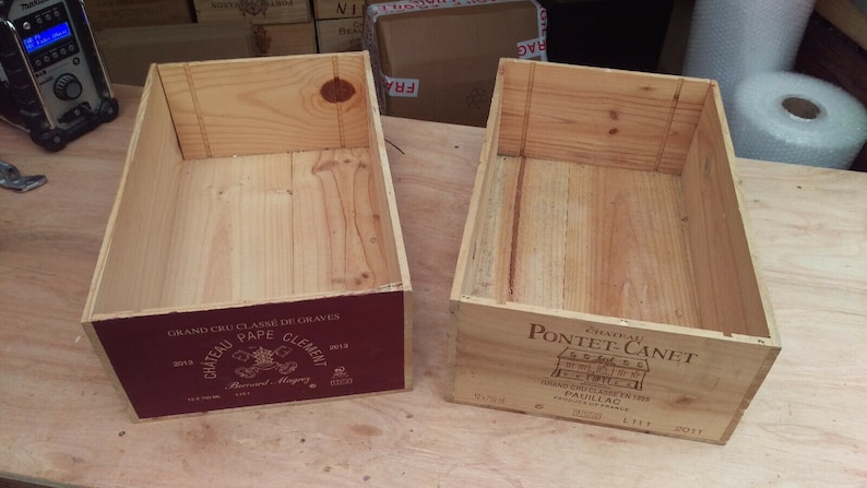 Pu&ograve; includere: Due cassette di legno per vino vuote. La cassetta a sinistra ha un'etichetta rossa con il testo "Grand Cru Classe de Graves Chateau Pape Clement 2012." La cassetta a destra ha un'etichetta bianca con il testo "Chateau Pontet-Canet Grand Cru Classe 1855 Pauillac 2011."