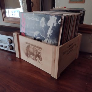 Op de afbeelding: Een lichtgekleurde houten krat gevuld met een verzameling vinylplaten. De voorkant van de krat bevat een gedrukte illustratie van een neushoorn en de tekst CAMPE DELLA SPINETTA BAROLO. De voorste plaat toont The Clash.