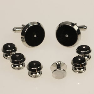 May include: A set of cufflinks and collar stays with black and silver accents. Each piece features a small, clear rhinestone in the center.