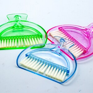 May include: Three small plastic dustpans with brushes. The dustpans are green, pink, and blue. Each dustpan has a handle and a brush attached to the bottom.