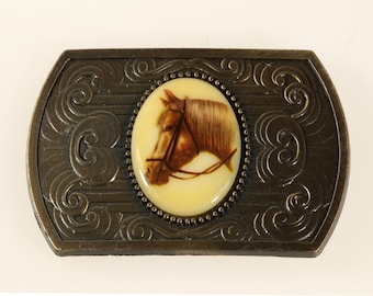 May include: A gold-toned belt buckle with an oval-shaped, cream-colored inlay featuring a brown horse head with a bridle.