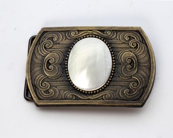 May include: A gold-toned belt buckle with an ornate design featuring a white mother-of-pearl inlay.