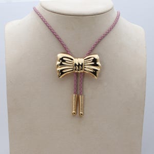 May include: A gold bow tie pendant on a pink braided leather bolo tie.