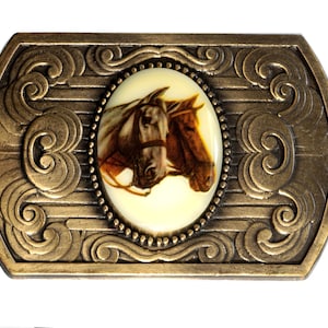 May include: A gold-toned belt buckle with an oval-shaped design featuring a white background and a picture of two brown horses' heads. The buckle has a decorative border with intricate swirls.