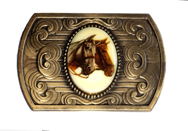 May include: A gold-toned belt buckle with an oval-shaped design featuring a white background and a picture of two brown horses' heads. The buckle has a decorative border with intricate swirls.