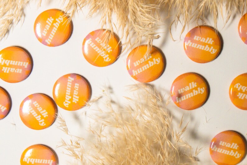 May include: A collection of round, orange and pink buttons with the text "walk humbly" in white lettering. The buttons are scattered on a white surface with dried beige grass.