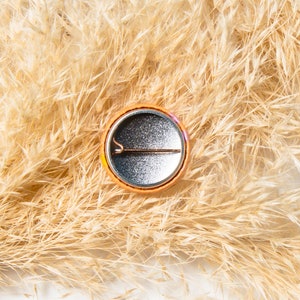 May include: A round, orange button with a silver back and a pin on the back. The button is sitting on a bed of dried beige grass.
