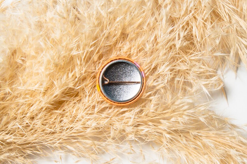 May include: A round, orange button with a silver back and a pin on the back. The button is sitting on a bed of dried beige grass.