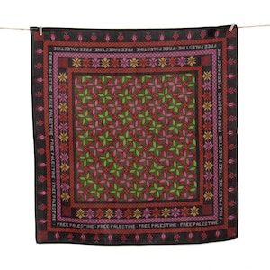 Pode incluir: Um lenço quadrado com um padrão geométrico detalhado em tons de verde, vermelho, rosa e amarelo. O lenço tem uma borda preta com o texto "FREE PALESTINE" repetido ao longo das bordas. O lenço está pendurado em um varal.