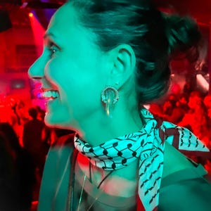 Könnte beinhalten: Eine Frau trägt silberne Ohrringe mit kreisförmigem Design und einen Schal mit einem schwarz-weißen Muster. Der Schal hat einen roten Rand. Der Hintergrund ist ein schwach beleuchteter Raum mit roter und grüner Beleuchtung.