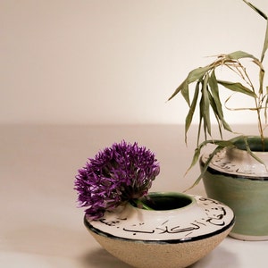 May include: A small, handcrafted ceramic vase with a purple flower and a green rim. The vase has Arabic script around the rim. A second, larger vase with green accents and bamboo is in the background. The vases are on a white surface.
