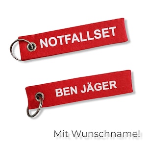 Könnte beinhalten: Zwei rote Stoffschlüsselanhänger mit weißem Text. Der obere Schlüsselanhänger sagt "NOTFALLSET" und der untere Schlüsselanhänger sagt "BEN JÄGER".
