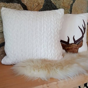 May include: Two white throw pillows, one with a cable knit texture and the other with a brown deer silhouette. The pillows are on a white faux fur rug.
