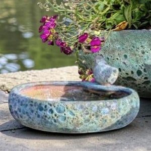Può includere: Tre fioriere in ceramica turchese di varie dimensioni, piene di fiori colorati. I fiori includono fiori arancioni, gialli, viola e rosa. Una piccola statuina di uccello decora la fioriera più piccola. Le fioriere sono posizionate su una superficie di pietra.
