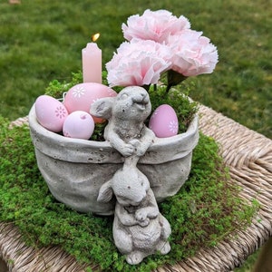 Pode incluir: Um vaso de concreto em forma de coelho segurando um coelho menor. O vaso está cheio de musgo, ovos de Páscoa rosa e cravos rosa.