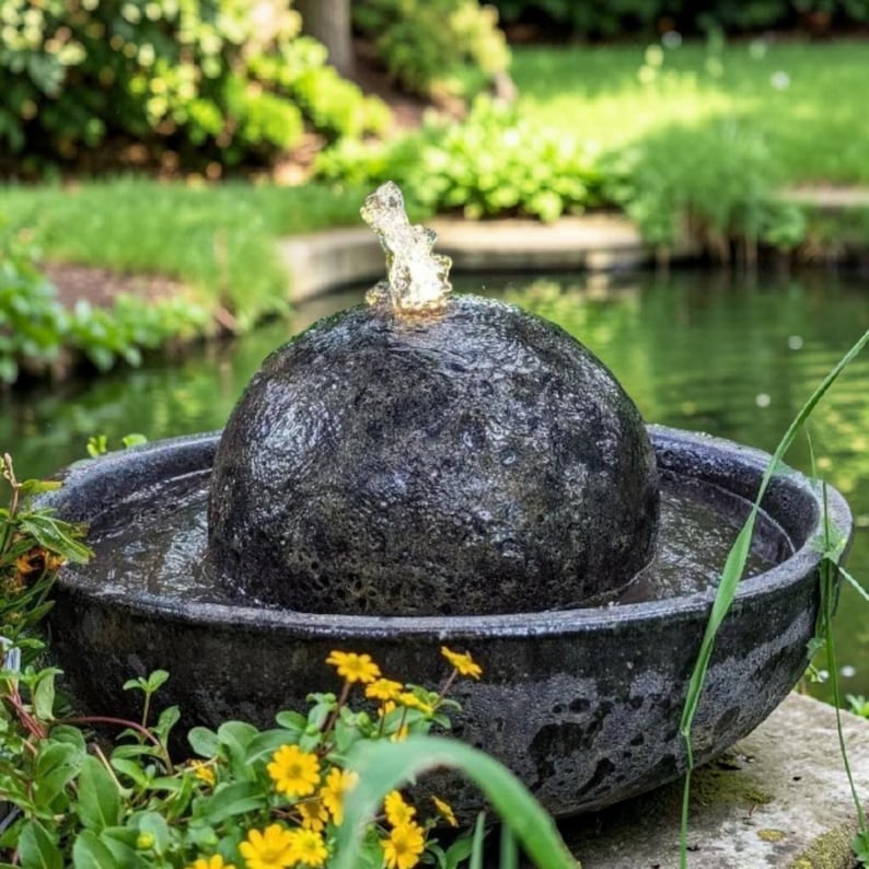 May include: A dark gray, textured stone water fountain with a spherical top and a wide, shallow basin. Water streams from the top, creating a small fountain. The fountain is surrounded by greenery and yellow flowers, set in a garden.
