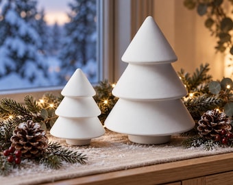 May include: Two matte white ceramic Christmas tree figurines with a smooth, tiered conical shape. The smaller tree stands to the left of the larger tree, both featuring a simple, clean design with a flat base, displayed as decorative objects.