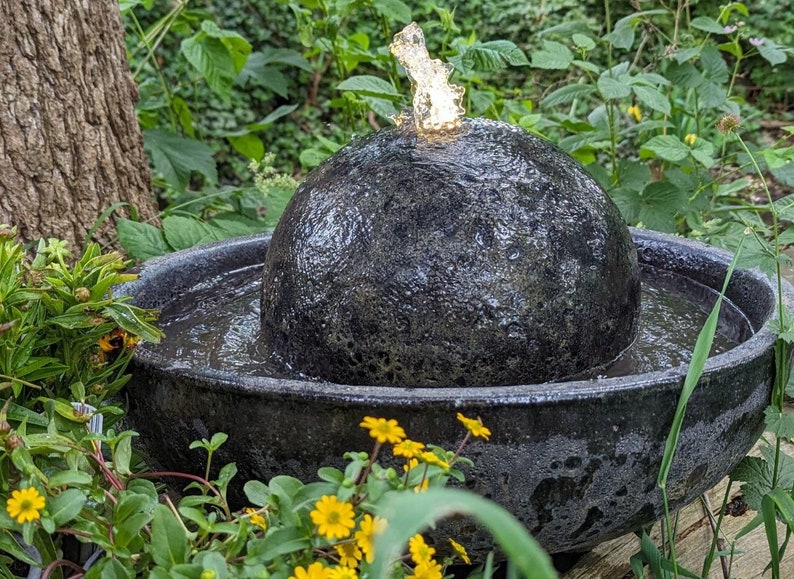 May include: A dark gray, spherical water fountain with water spouting from the top. The fountain sits in a round, dark gray basin. Yellow flowers and green foliage surround the fountain, creating a natural, outdoor aesthetic.